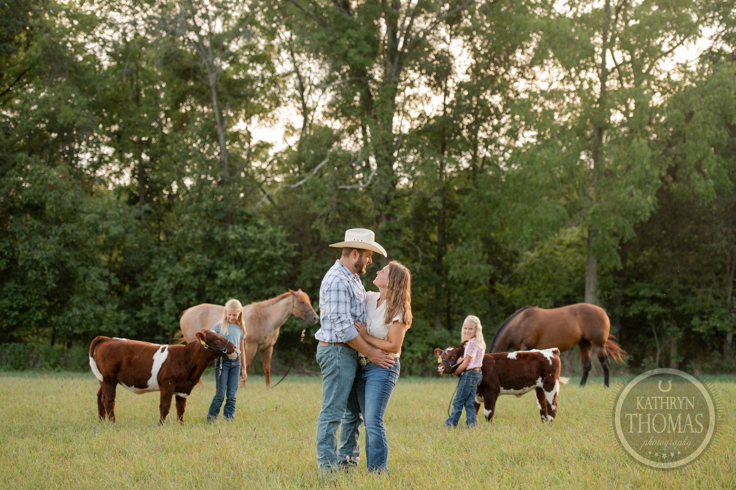 A group stands together in a grassy field with horses and calves during golden hour.