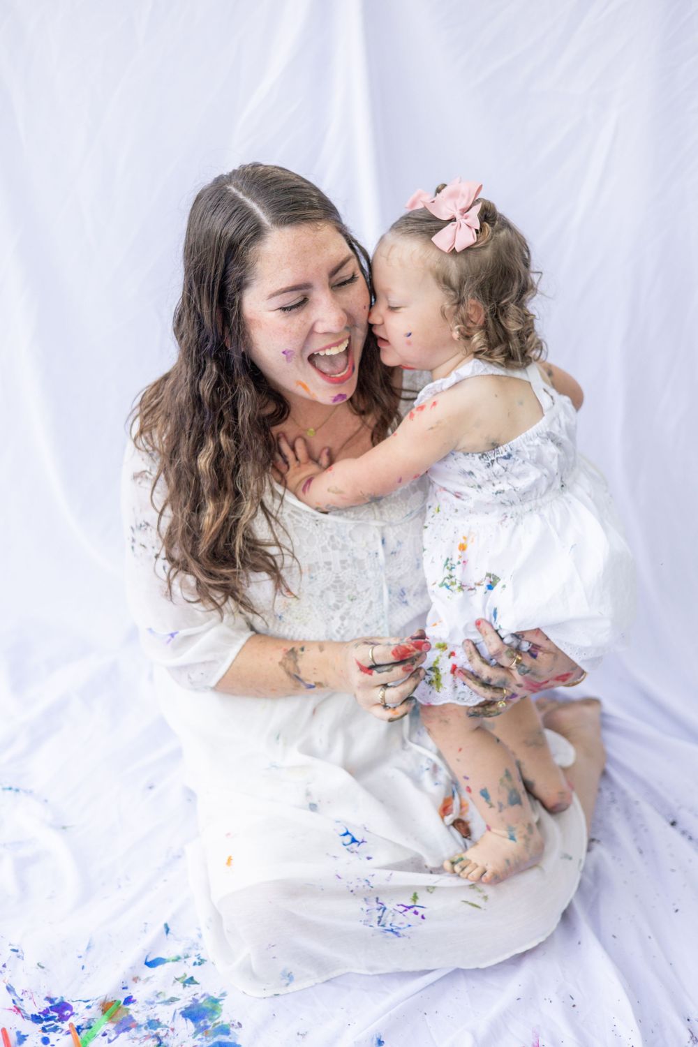 A mother and toddler in white dresses share a joyful moment together surrounded by blue paint splatters on white fabric.