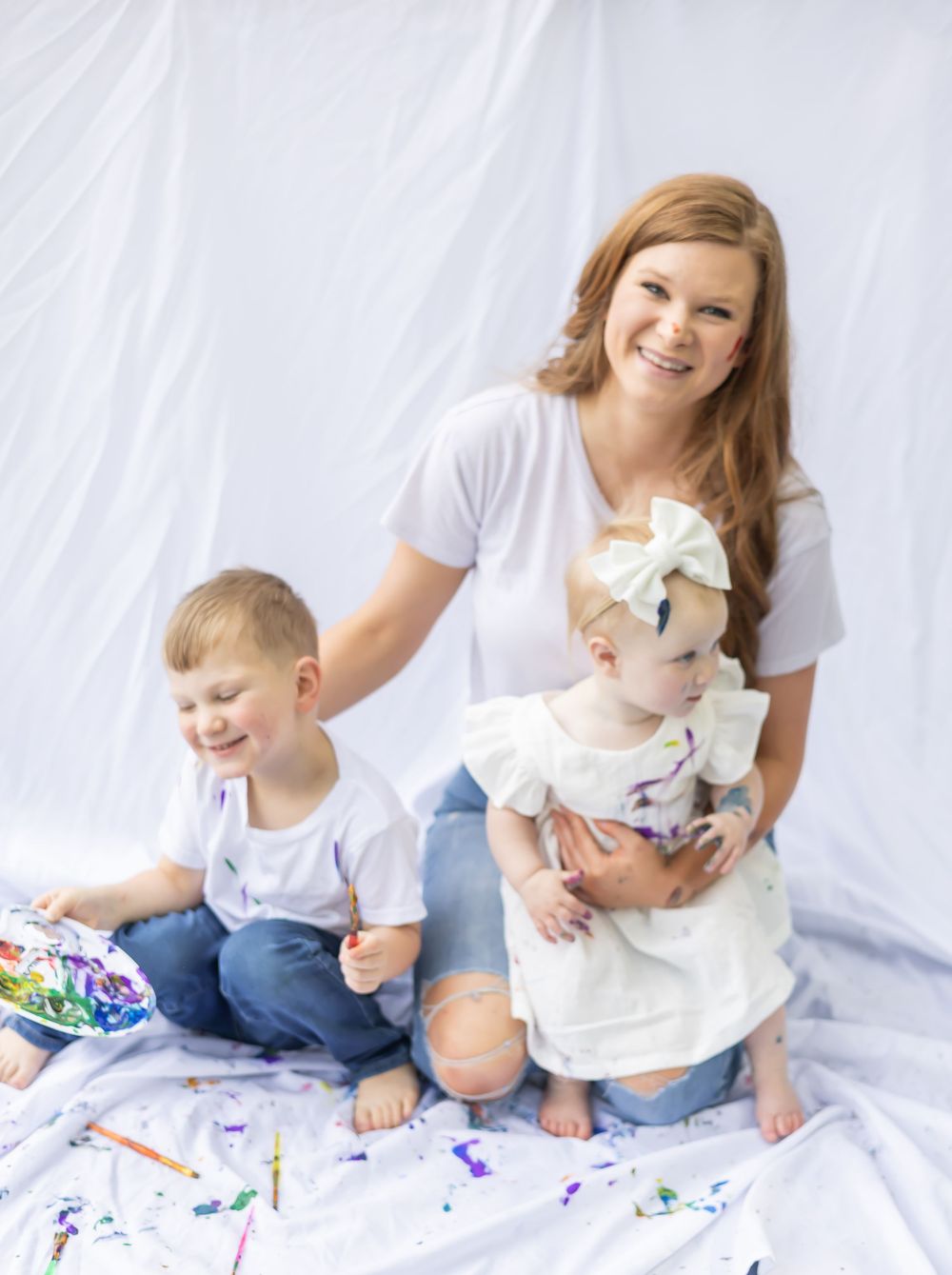 Family enjoys creative time together on white sheet with colorful paint and art supplies in bright setting.