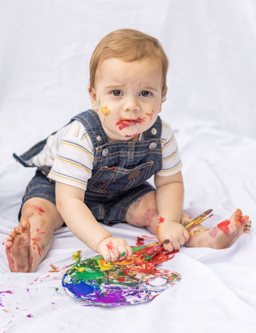 Baby in denim overalls sitting on white sheet and finger painting with colorful paint, messy with paint on face.