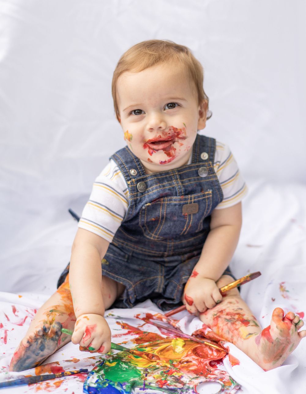 Baby in denim overalls creates colorful mess while finger painting with bright rainbow paints on white background.
