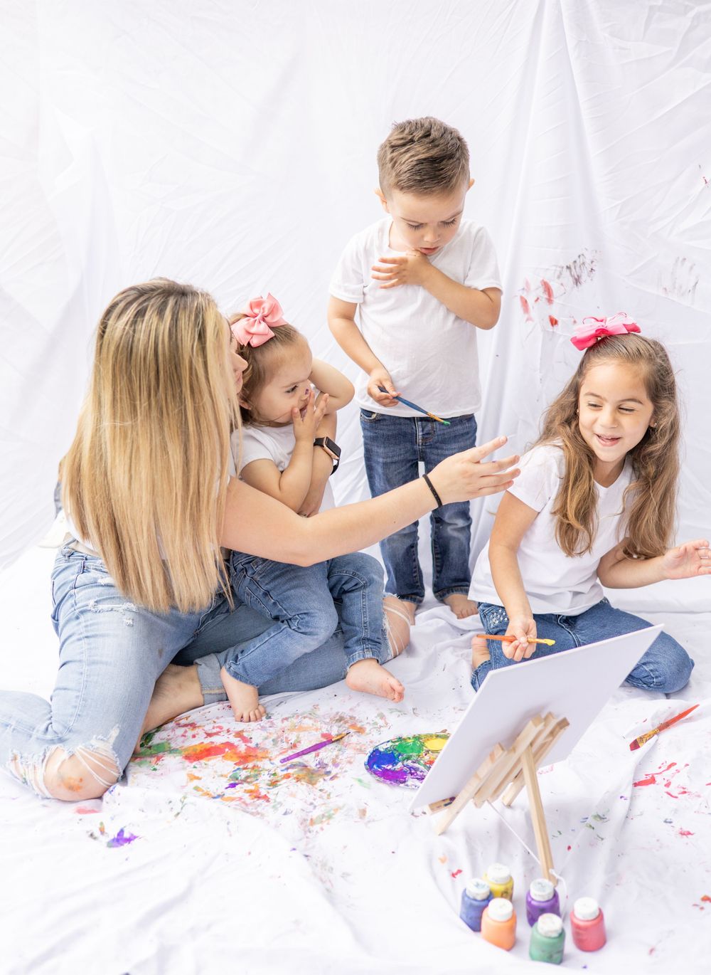 Small family sitting on white background doing colorful finger painting together on paper while wearing casual clothing.