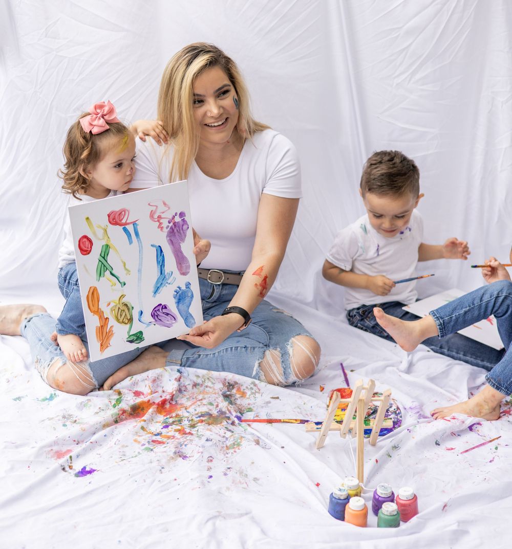 Children and adult sitting on white backdrop with art supplies and colorful paintings during creative playtime.
