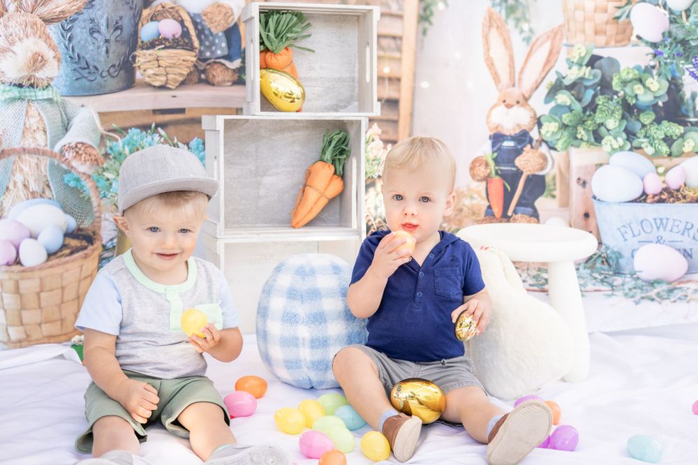 Two toddlers sit together in an Easter-themed studio setup with carrots, eggs, and baskets in soft pastel colors.