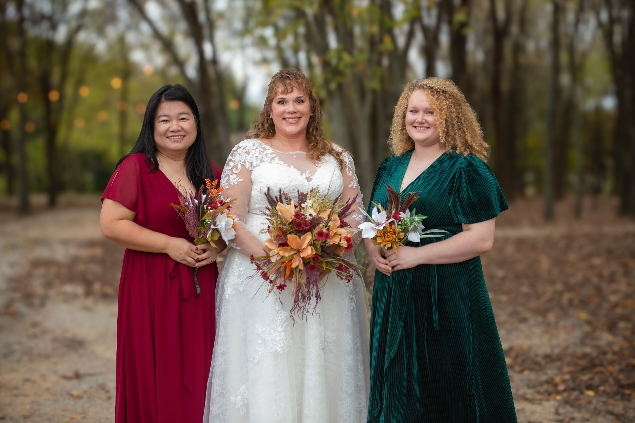 A bridal party poses together wearing burgundy and emerald dresses in a wooded setting.
