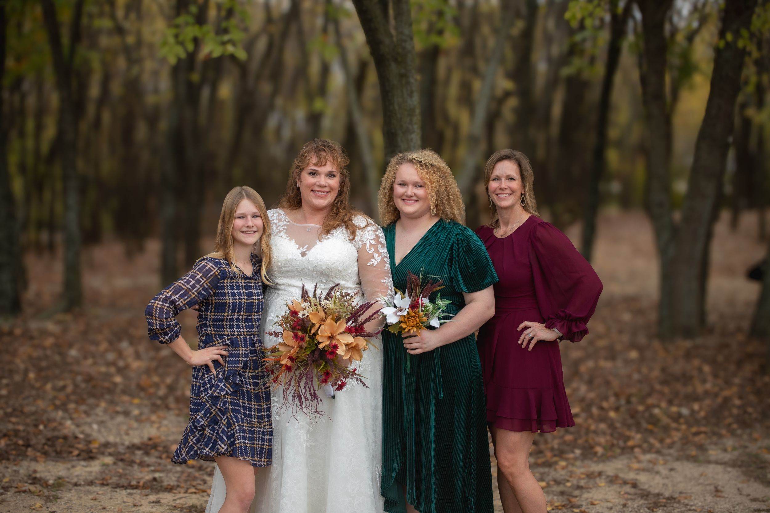 A group poses for a wedding photo in casual attire among fall trees.