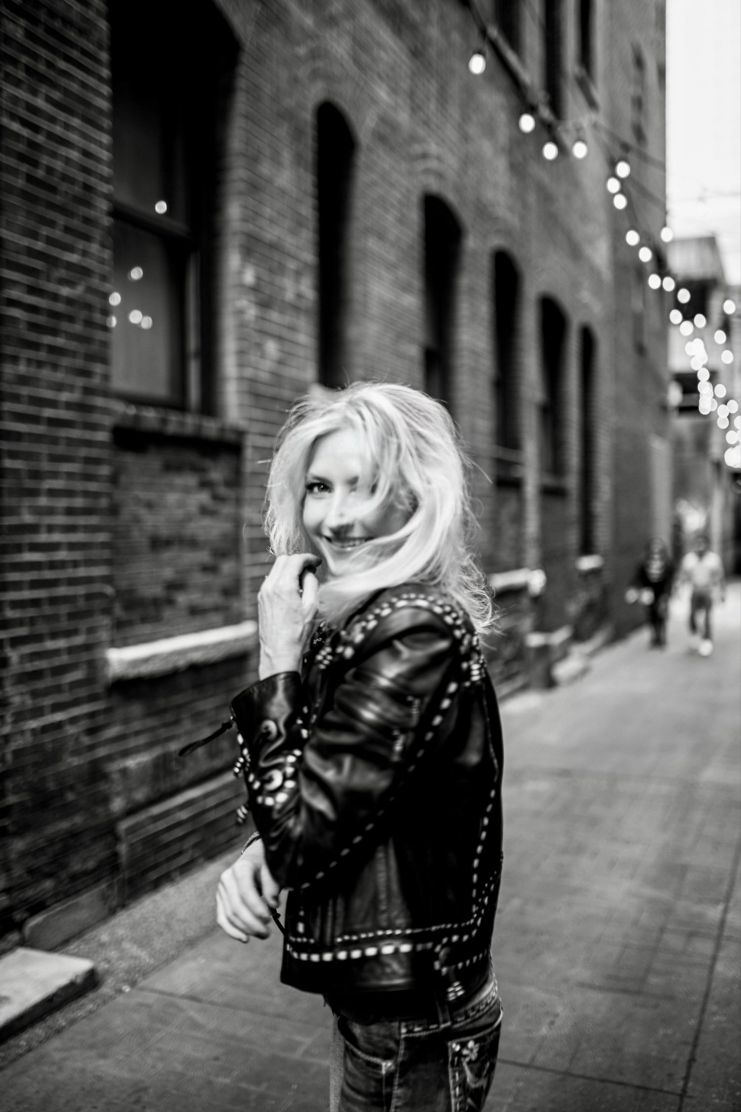 Artistic black and white street portrait with string lights and brick wall backdrop showing person in leather jacket.