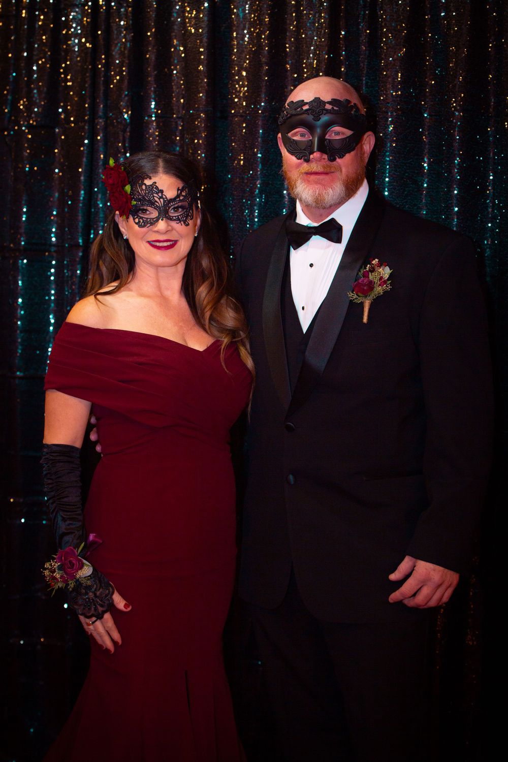 Formal couple in burgundy dress and black tuxedo pose at masquerade ball with glittering backdrop.