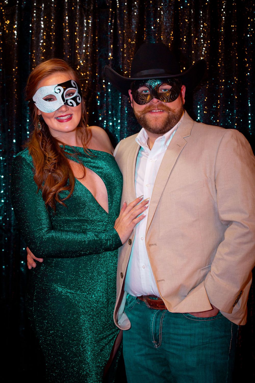 Couple poses at masquerade party with sparkly backdrop, wearing emerald dress and tan suit.