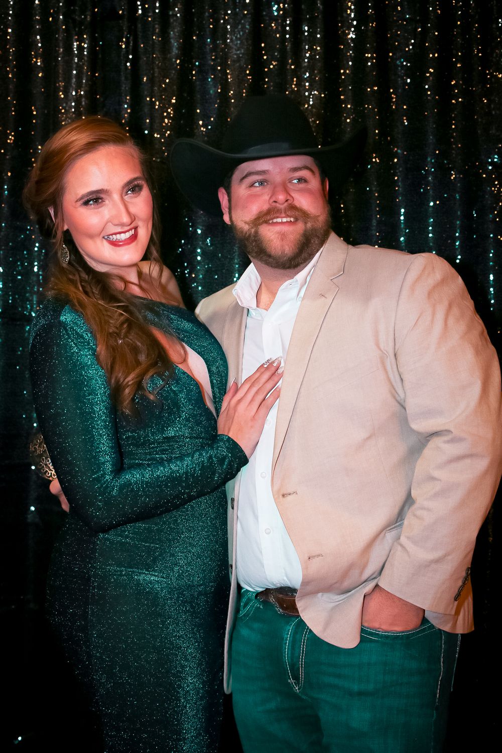 Glamorous couple in formal attire poses playfully against a sparkly backdrop at masked event.
