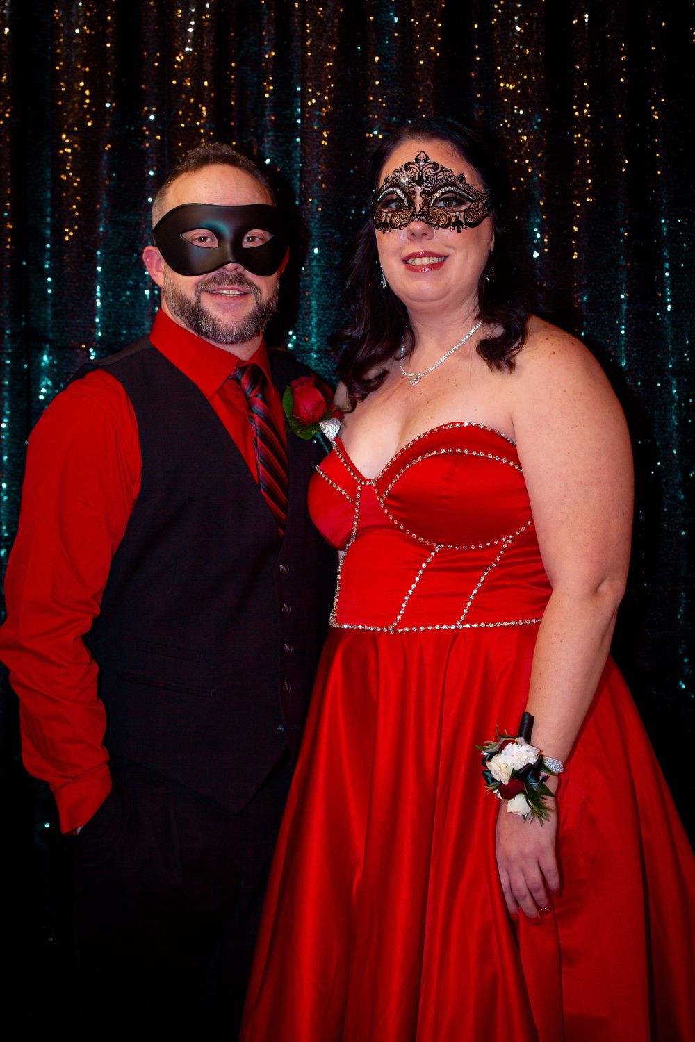 Couple in matching red and black attire poses at masked ball with sparkling curtain backdrop.