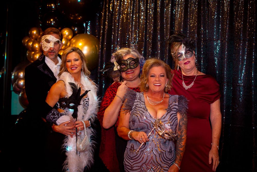 Masquerade ball attendees in formal evening wear pose against sparkly curtain background.