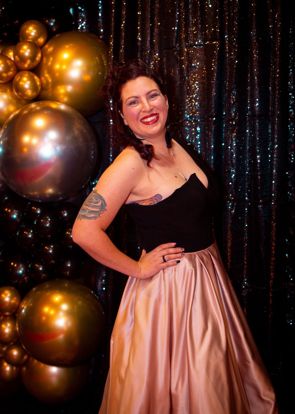 A person in a pink formal dress poses against a sparkly backdrop with gold balloons.