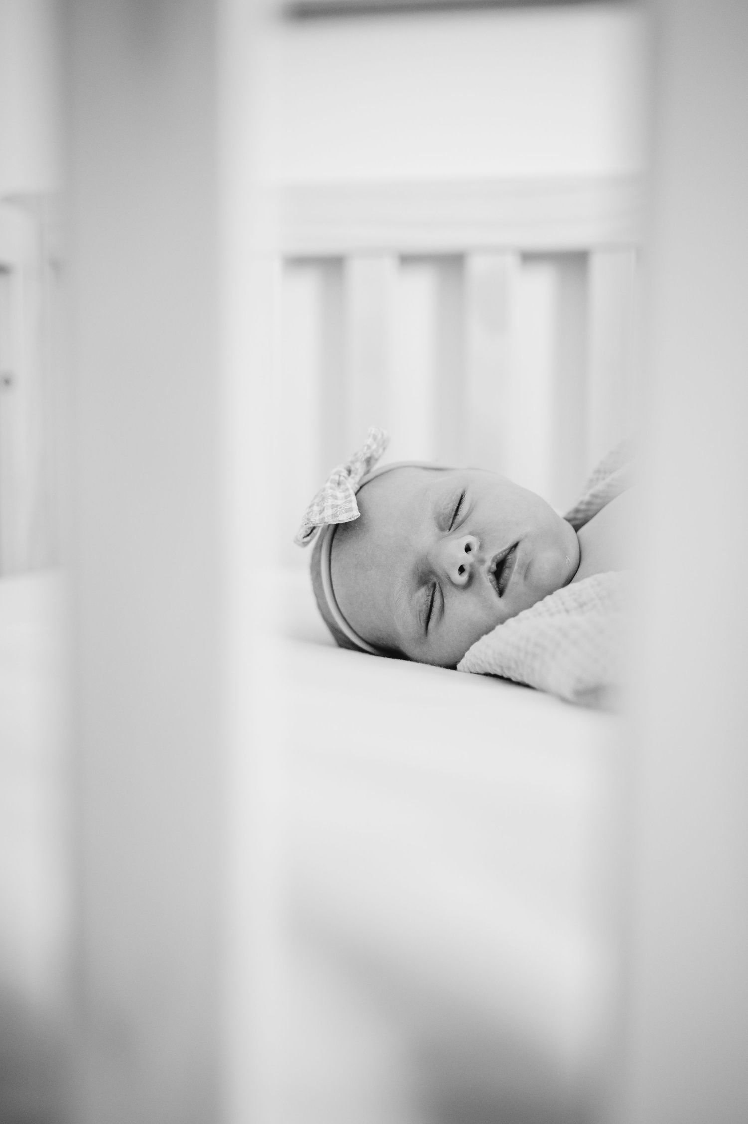 Black and white photo of a tiny infant resting peacefully in a white crib, viewed through the slats.