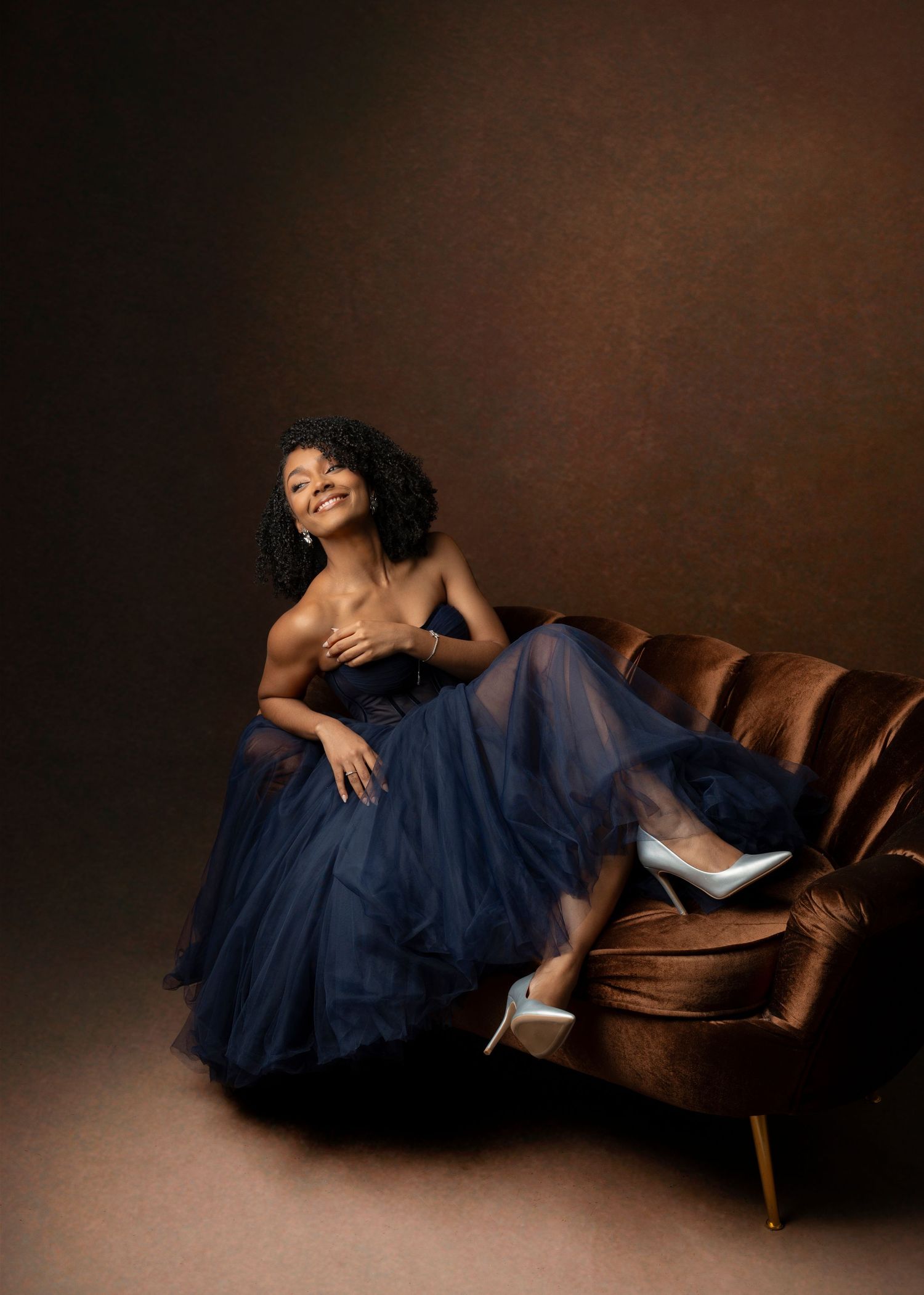 Artistic portrait featuring a woman in a flowing navy dress lounging on a mid-century leather sofa.