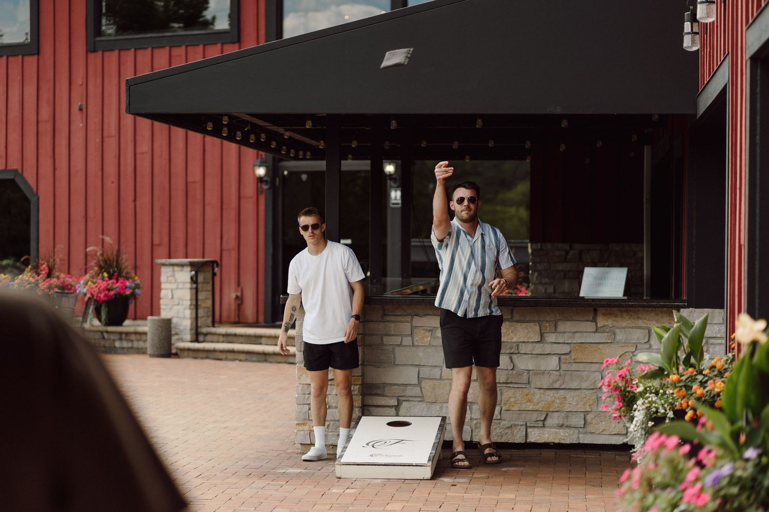 Two friends playing cornhole outside a red barn building on a sunny day.
