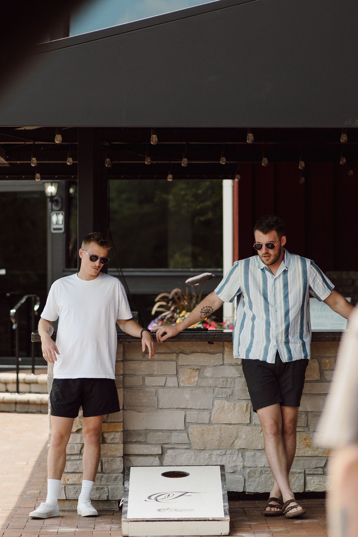 Two people playing cornhole game on stone patio near red building.