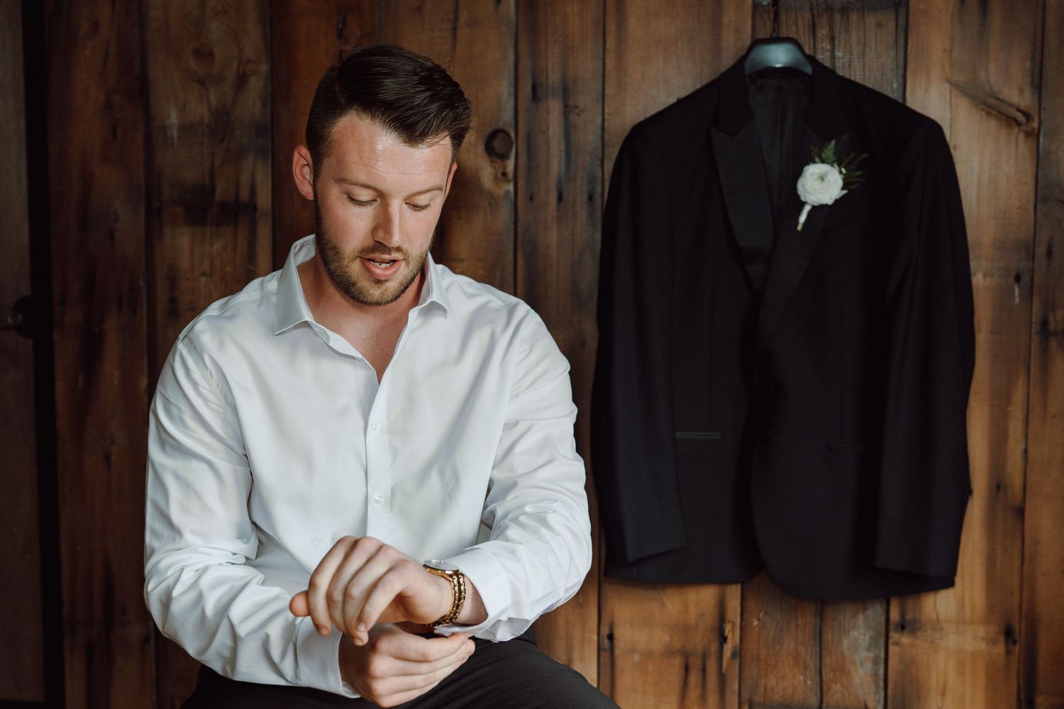 Formal black suit jacket with boutonniere hangs while person adjusts cuffs in rustic wood room.
