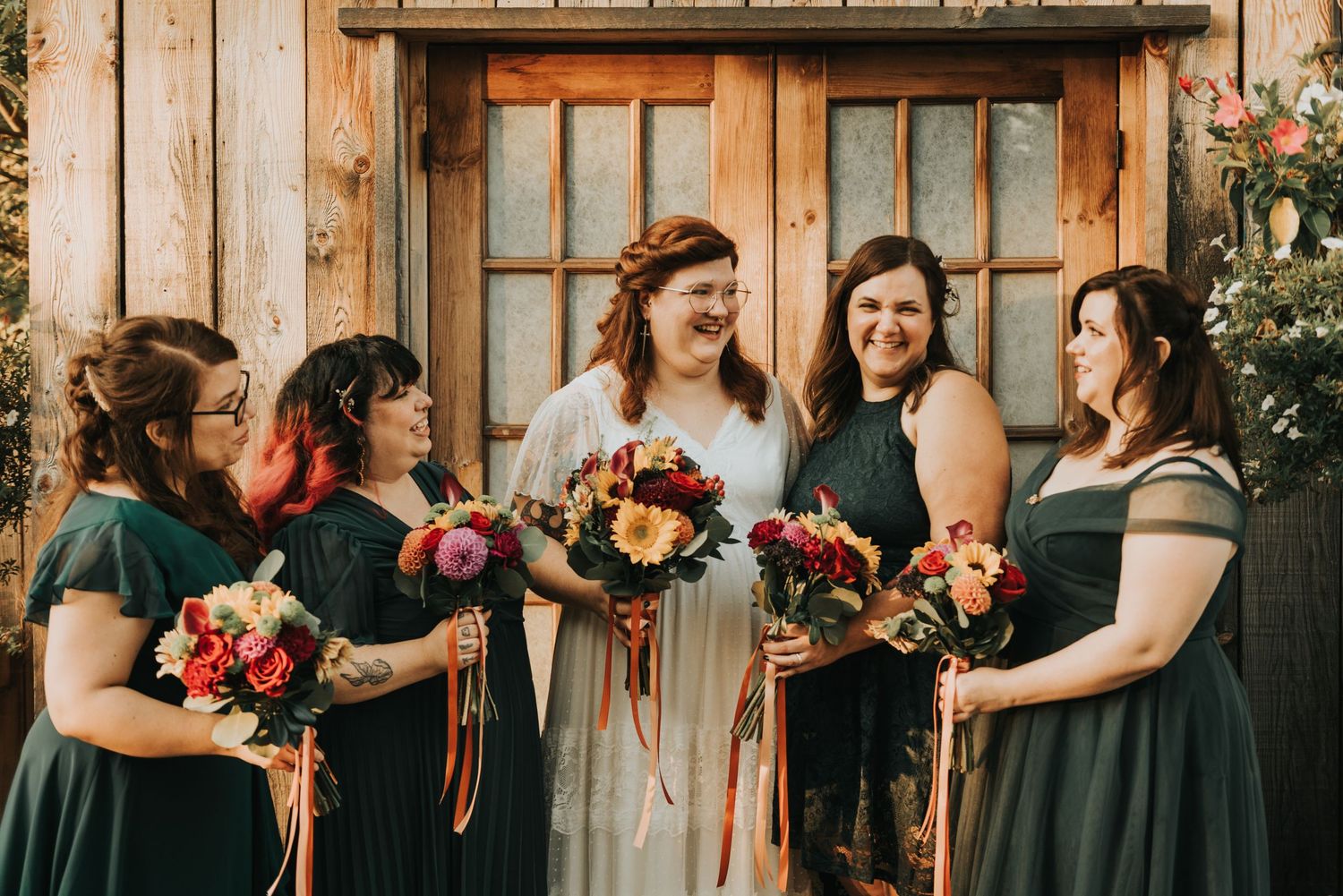 Wedding party stands outside wooden door wearing green dresses and holding autumn-colored bouquets.