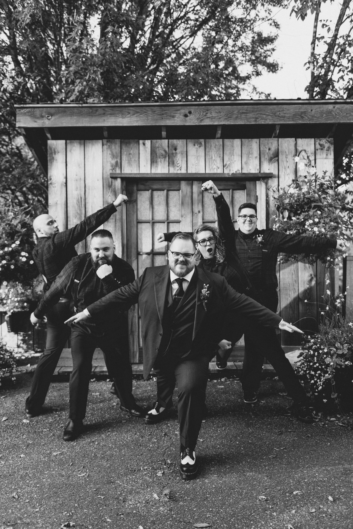 A black and white vintage photograph shows a group of men in suits striking dramatic poses outside a wooden shed.