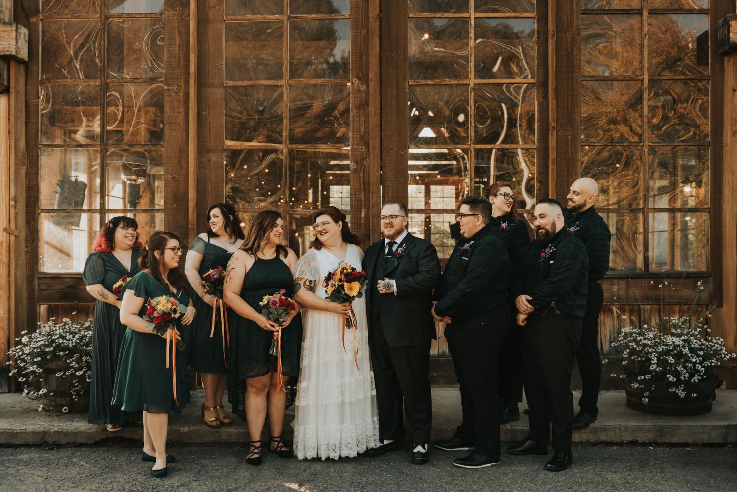 Wedding party poses together in front of rustic barn venue with natural wooden backdrop and floral arrangements.
