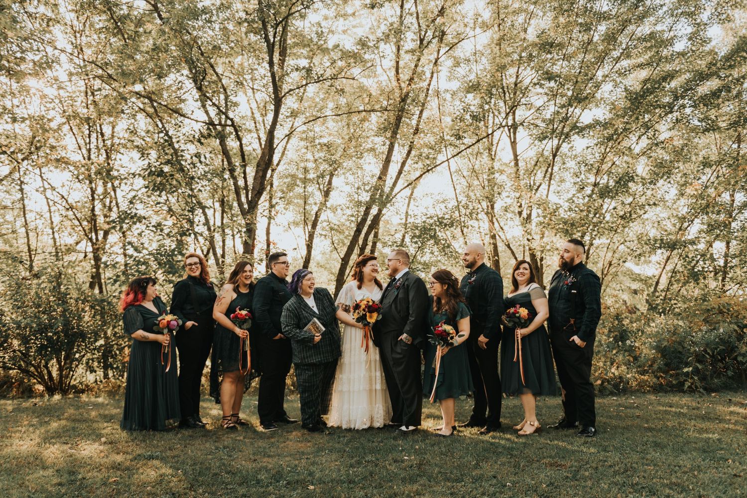 Wedding party in black attire poses together in sunlit wooded outdoor setting during golden hour.
