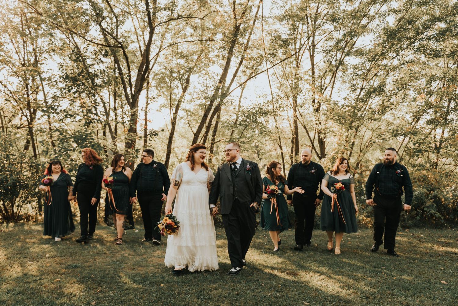 Wedding group gathers in casual poses among trees on sunny autumn afternoon.