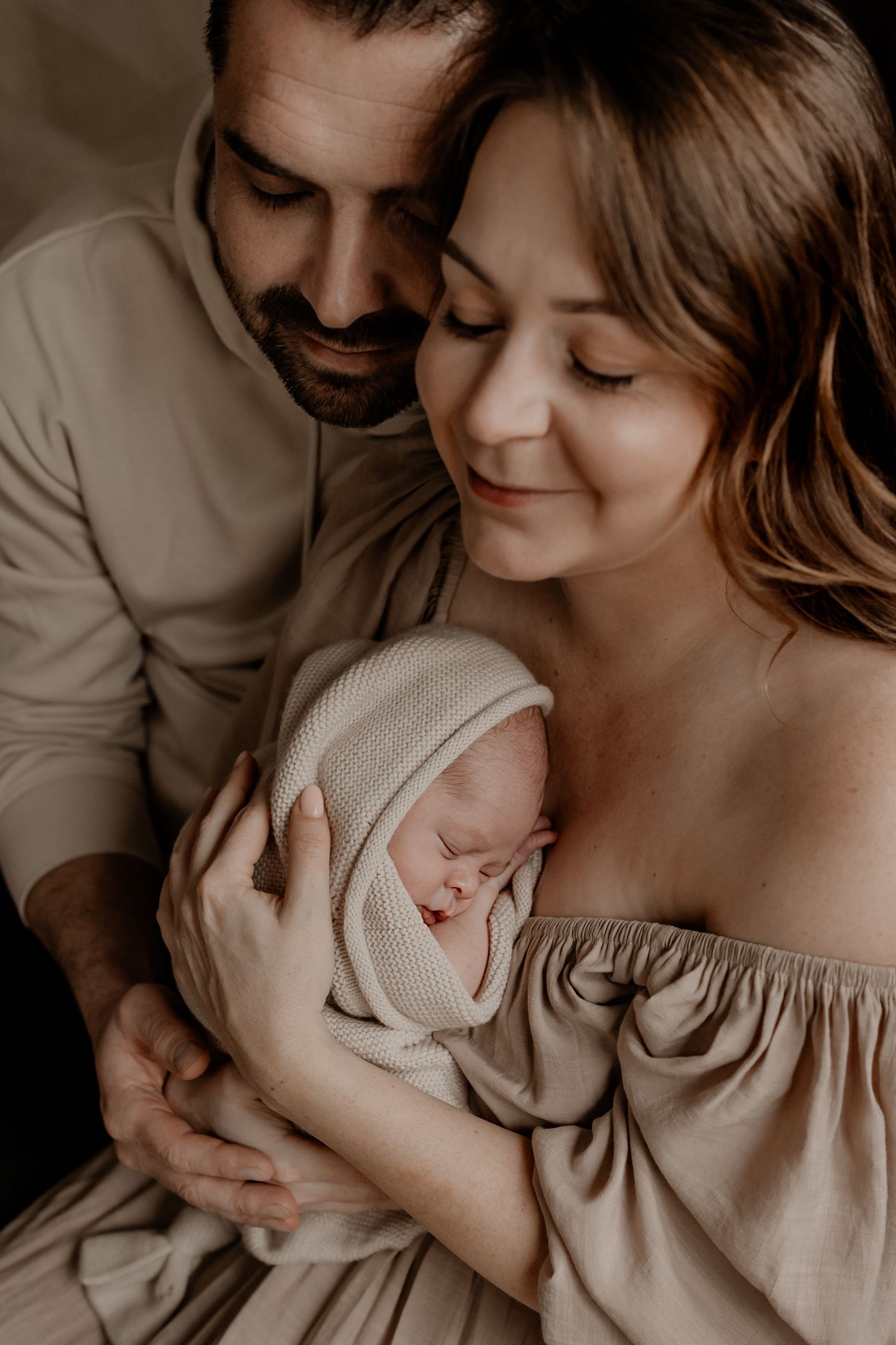 Intiem familieportret in zachte beige tinten, waarop ouders hun liefdevol gewikkelde pasgeboren baby teder omarmen.