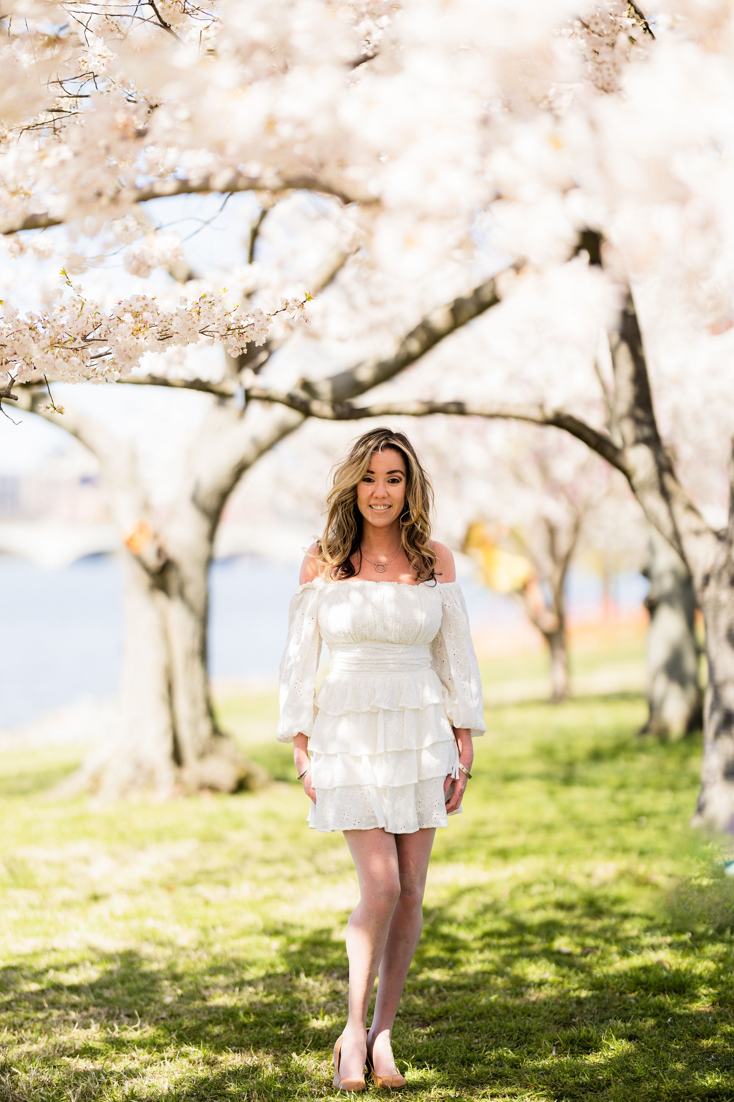 A figure in a white lace dress stands barefoot in a springtime garden beneath blooming cherry blossom trees.