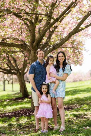 Family stands together under blooming pink cherry blossom trees in a spring garden setting.