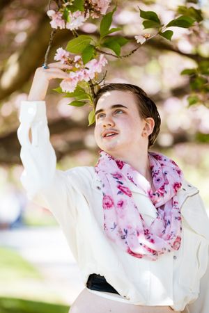 A person in white wearing a pink floral scarf gazes up at blooming cherry blossoms in spring.