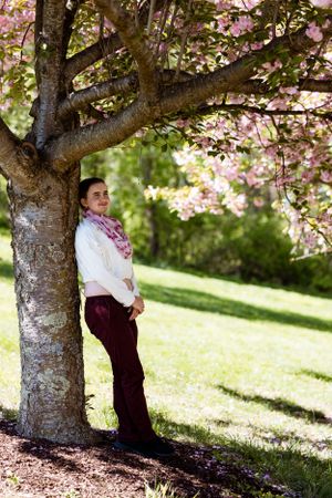 A person stands beneath a blooming cherry blossom tree on a sunny spring day in a grassy park setting.
