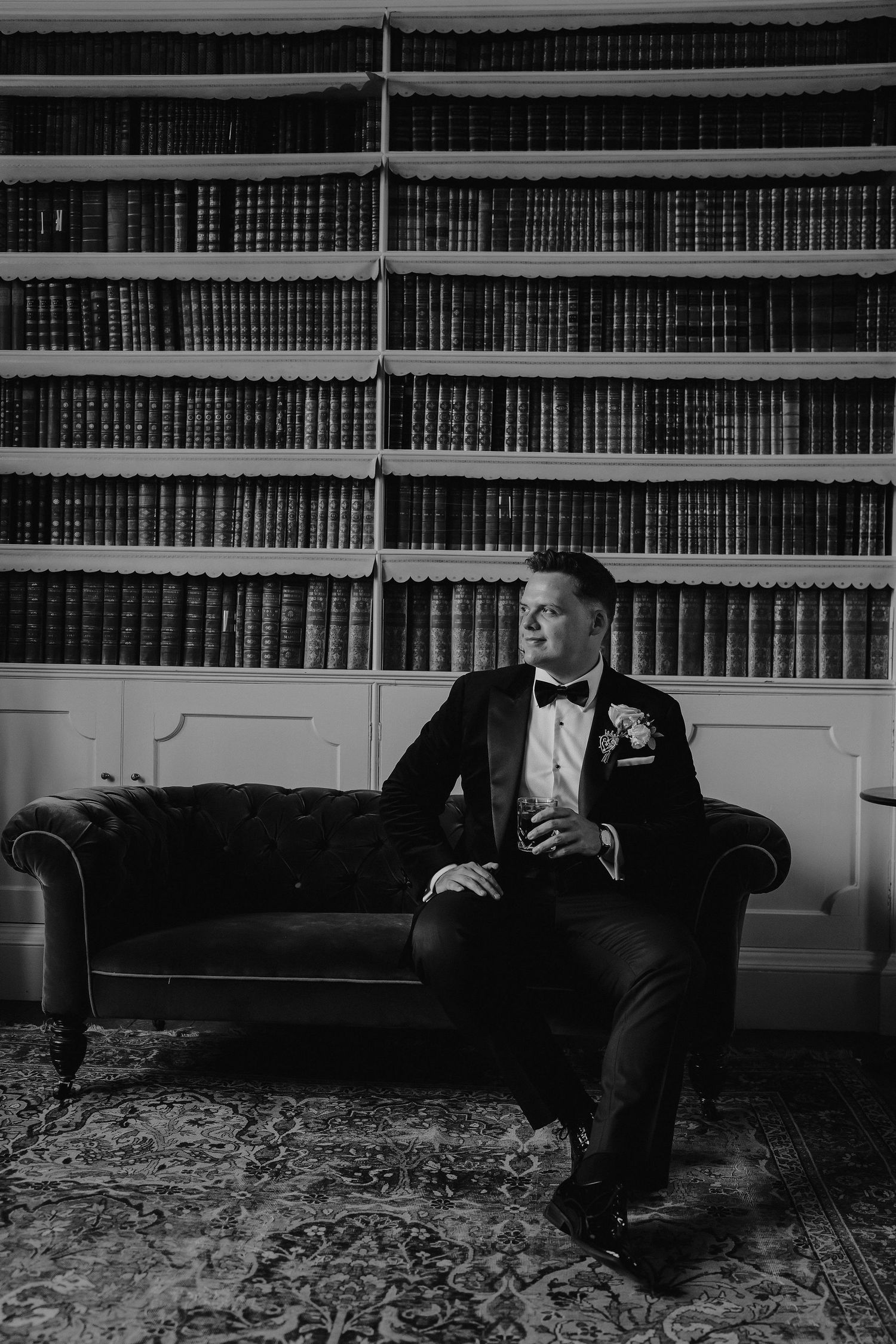 groom sat on sofa with a tumbler of whiskey in his hand in the library at Iscoyd park