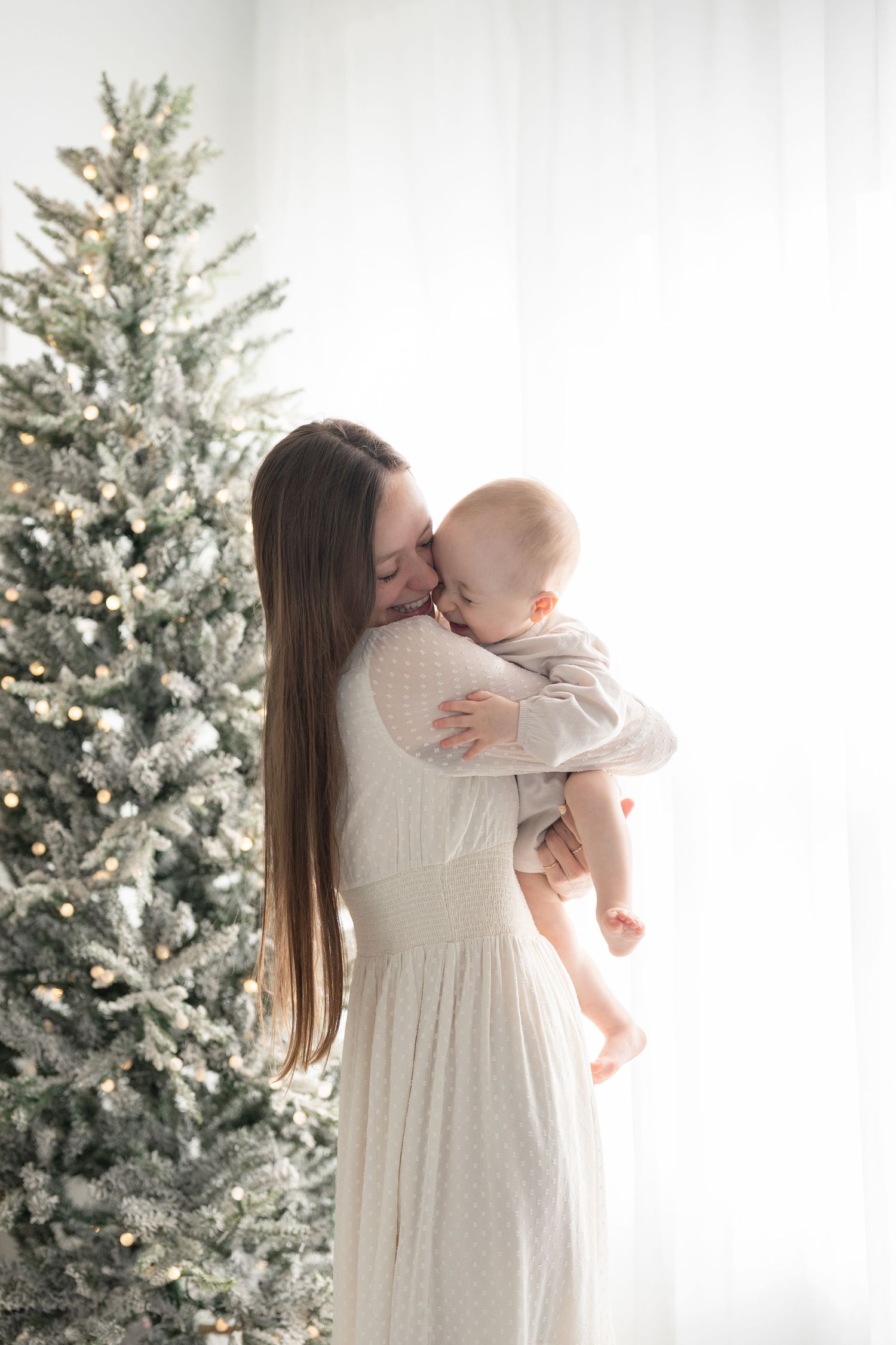 Portrait fine art de maman et bébé riant l'un contre l'autre devant un sapin enneigé et illuminé lors d'une séance photo de Noël au studio