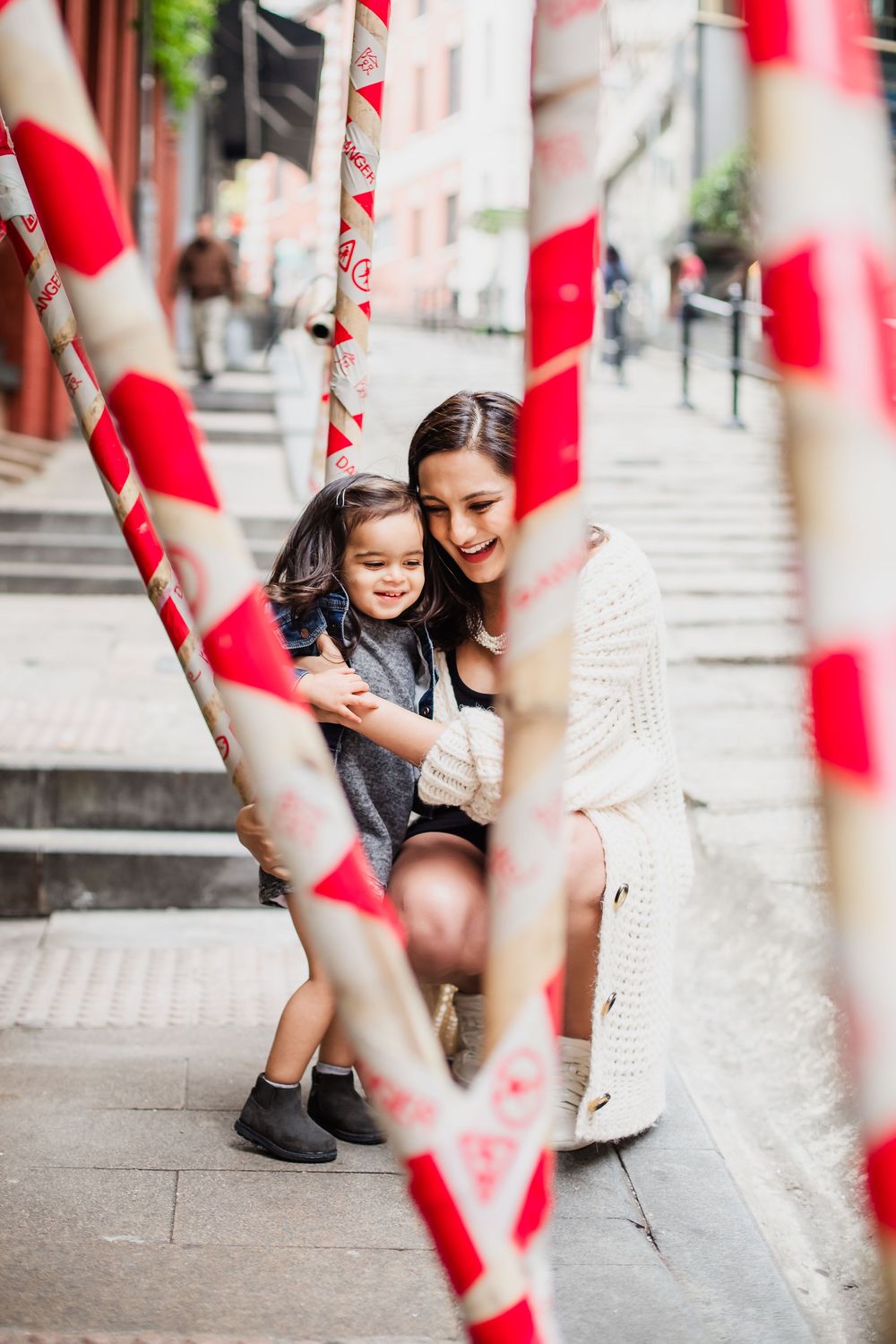Playful mother and daughter sit on concrete steps behind red and white striped barriers in an urban setting.