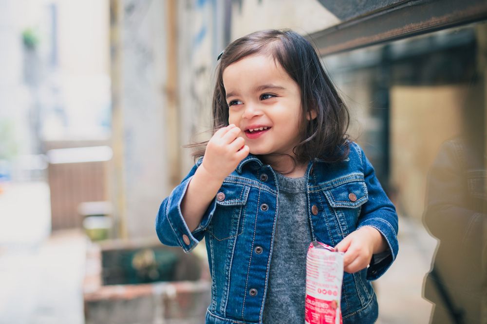 A happy child in a denim jacket holds a pink beverage container while standing outdoors.