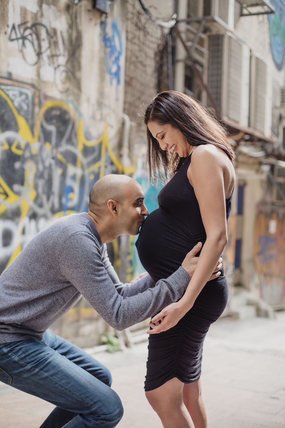 A tender pregnancy moment captured against an urban graffiti wall with black dress and gray sweater.