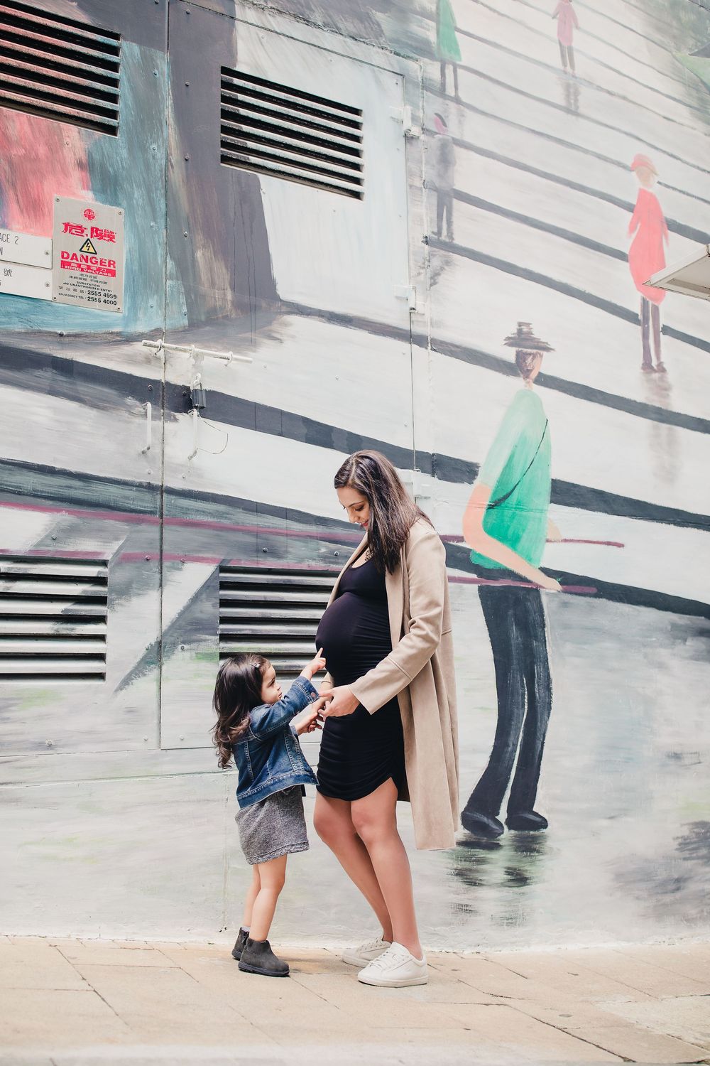 A person wearing a tan coat bends down near a wall while another person stands behind them.