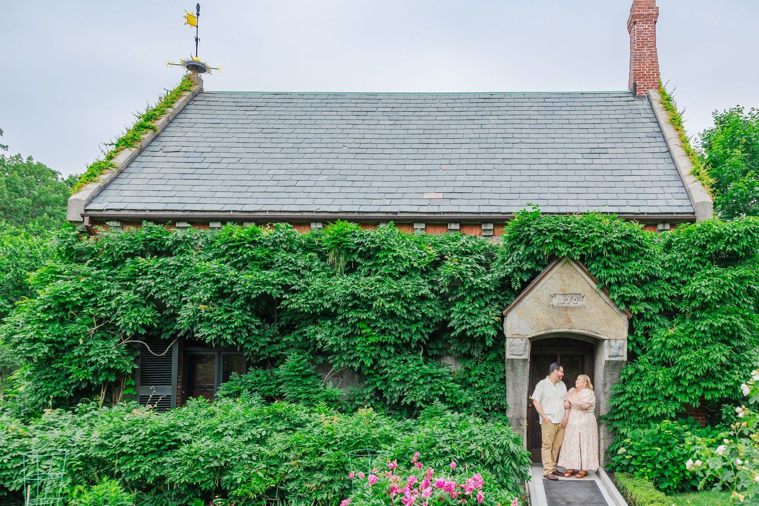 A cottage with gray slate roof is covered in lush green vines and features a sheltered entranceway with pink flowers.
