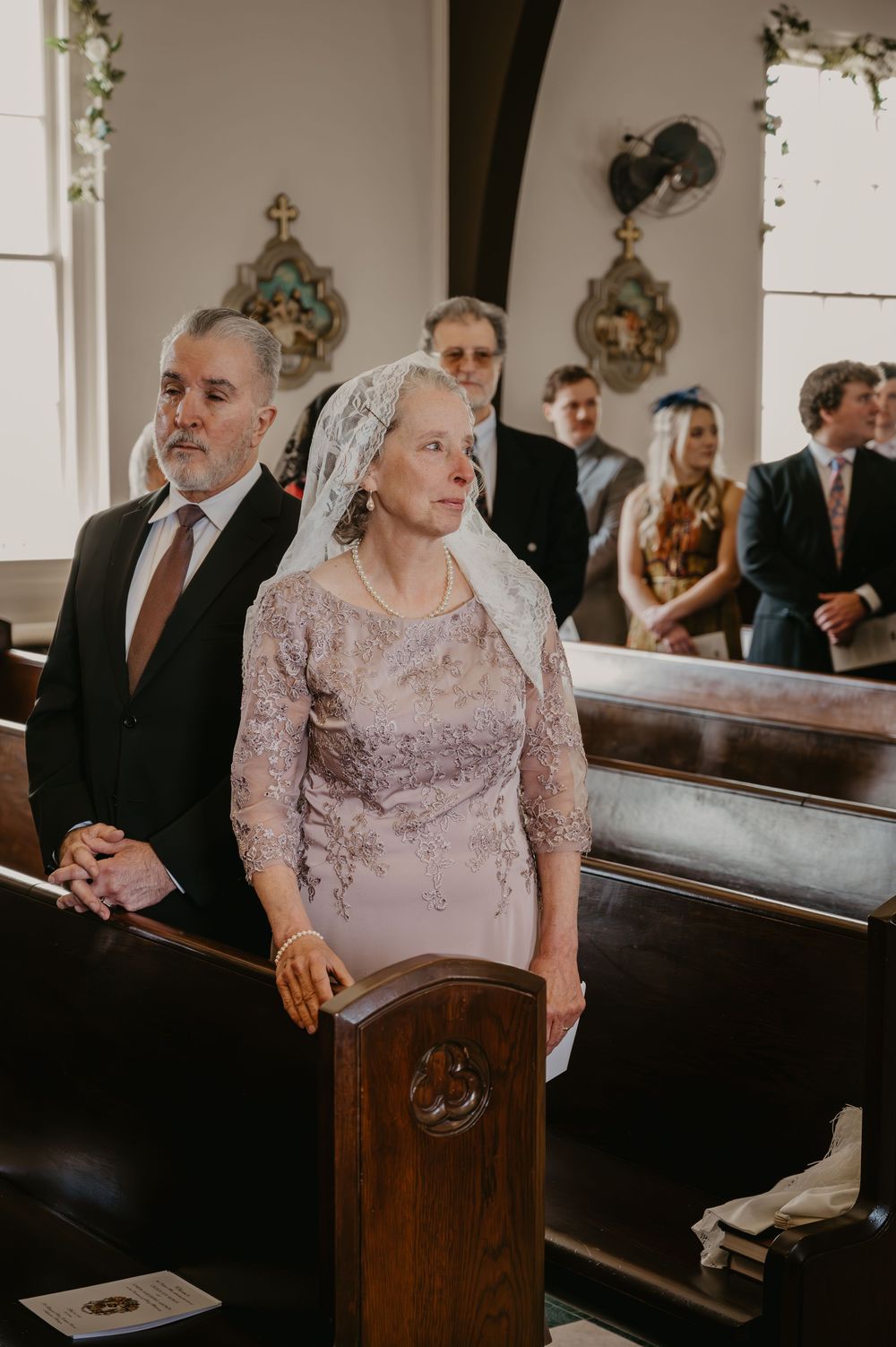 Wedding guests seated in church pews, woman in lace dress wearing traditional chapel veil during ceremony.