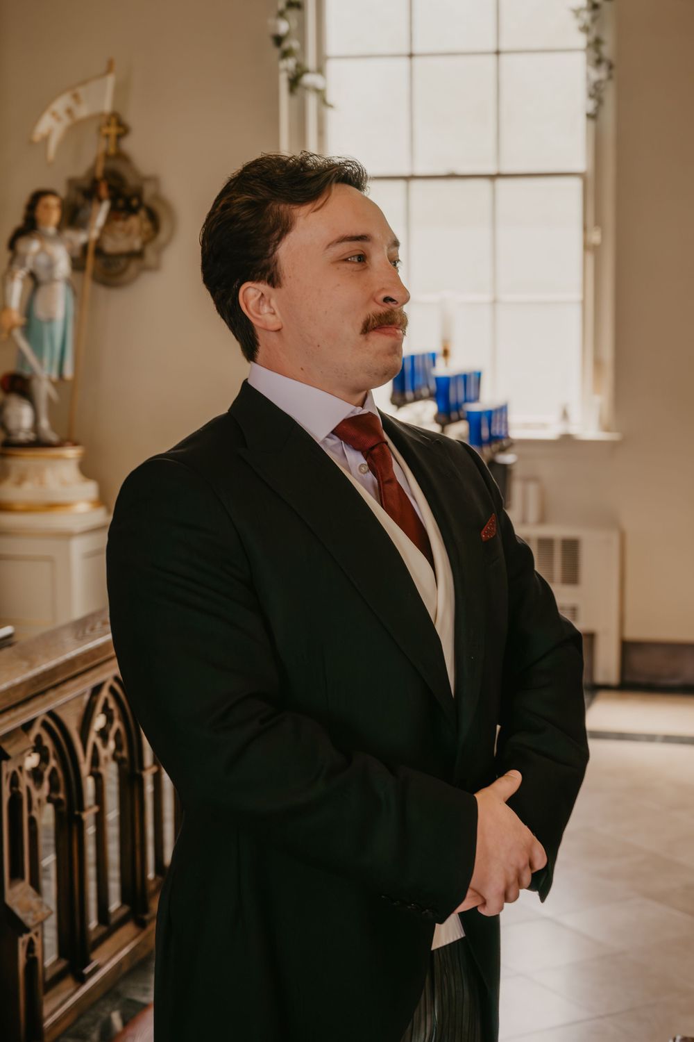 Elegant groom in black suit with red tie standing in historic church during wedding ceremony.