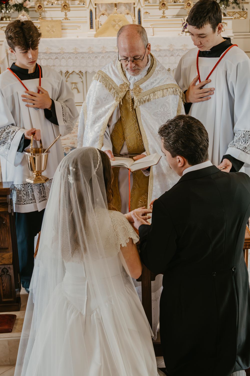 Bride and groom exchanging rings before priest at ornate Catholic church altar during wedding ceremony.