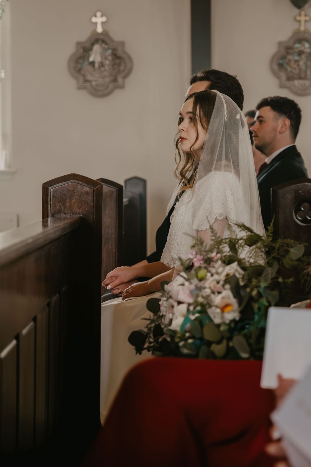 Beautiful bride in lace veil sitting in church pew holding elegant floral bouquet during wedding.