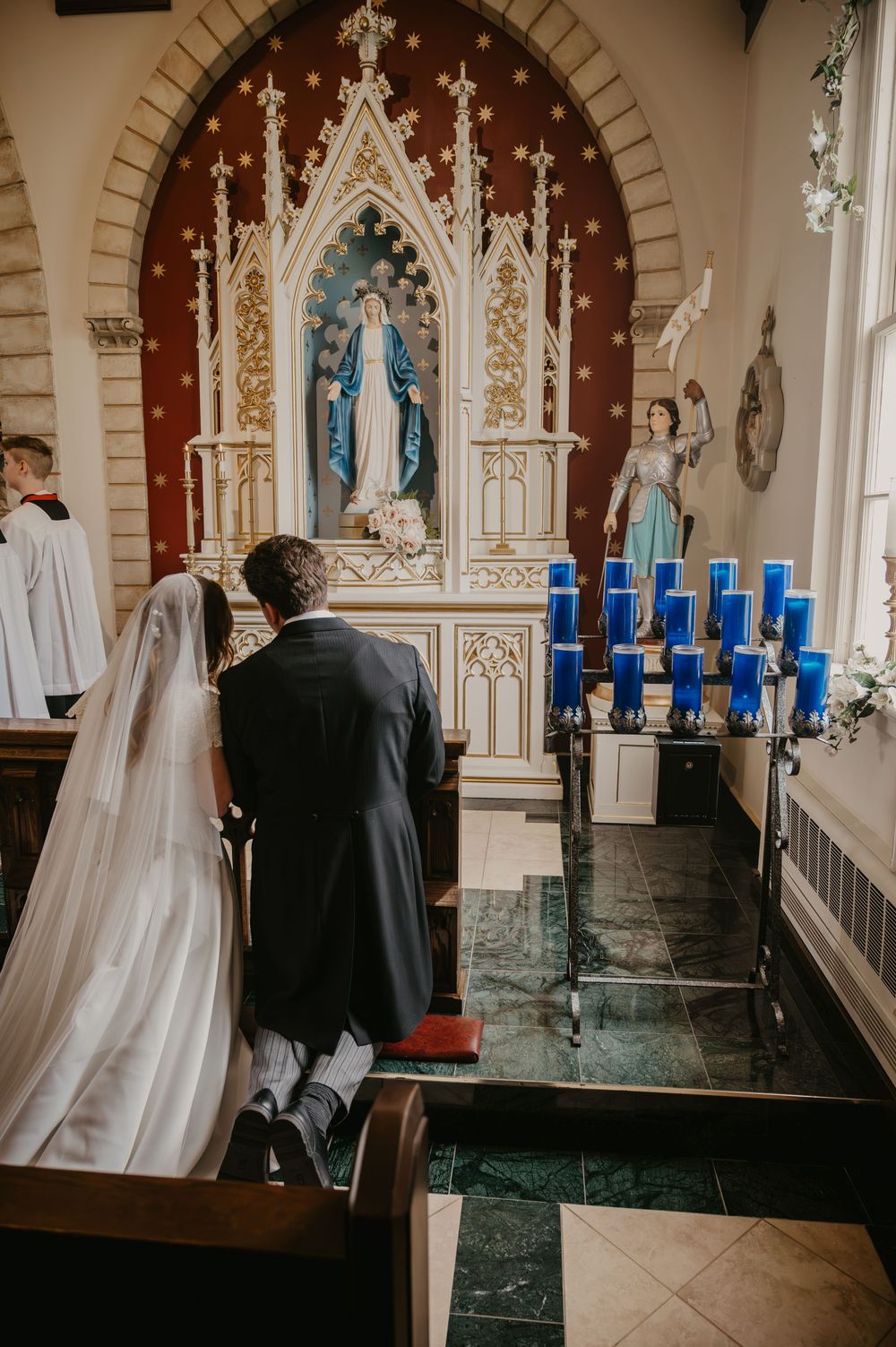 Bride and groom kneeling before Virgin Mary altar with blue candles during Catholic wedding ceremony.