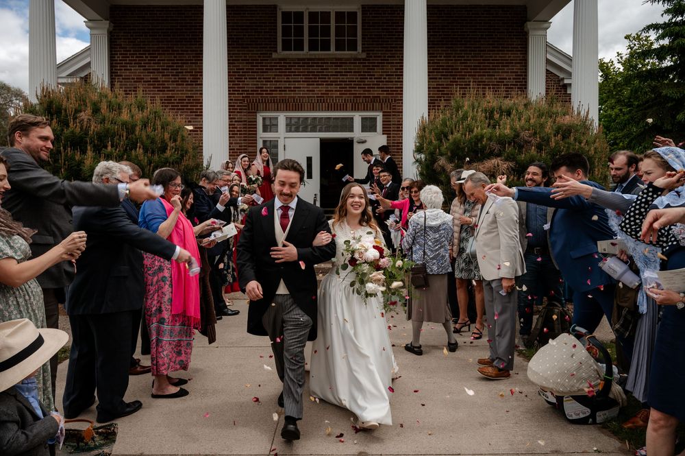 Joyful bride and groom exit brick church amid confetti shower from smiling wedding guests lining the path.