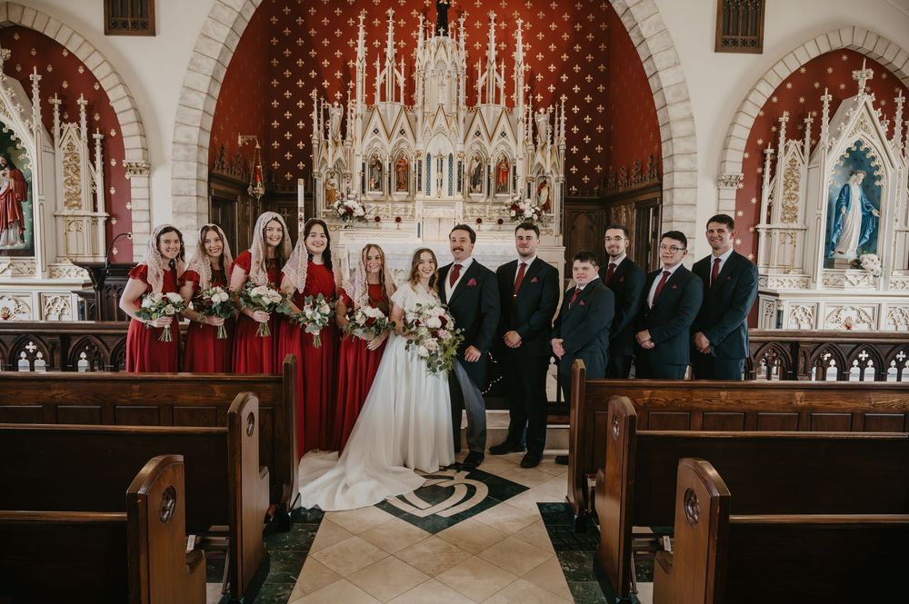 Wedding party poses in ornate red and white Gothic church with bridesmaids in burgundy gowns.