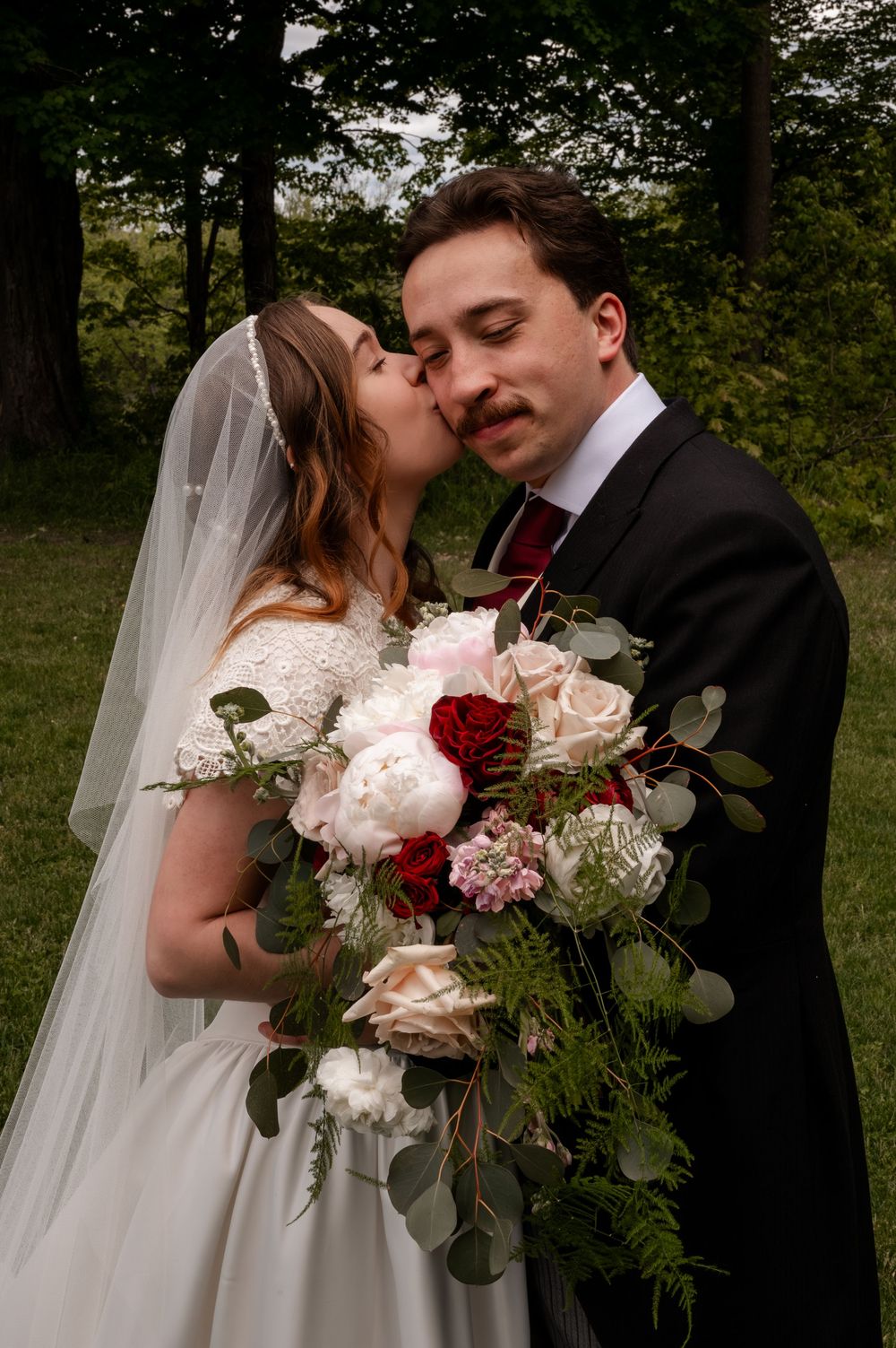 Bride kisses groom on cheek while holding lush bouquet of red roses and white peonies in garden setting.