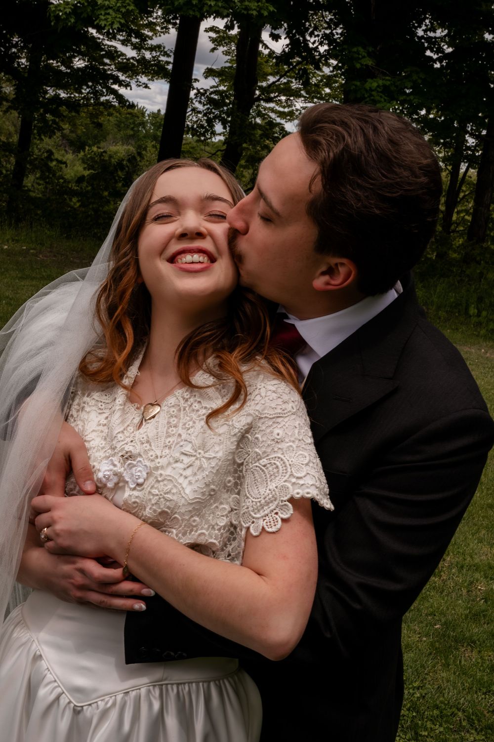 Groom kisses smiling bride on cheek while embracing her outdoors among lush green trees.