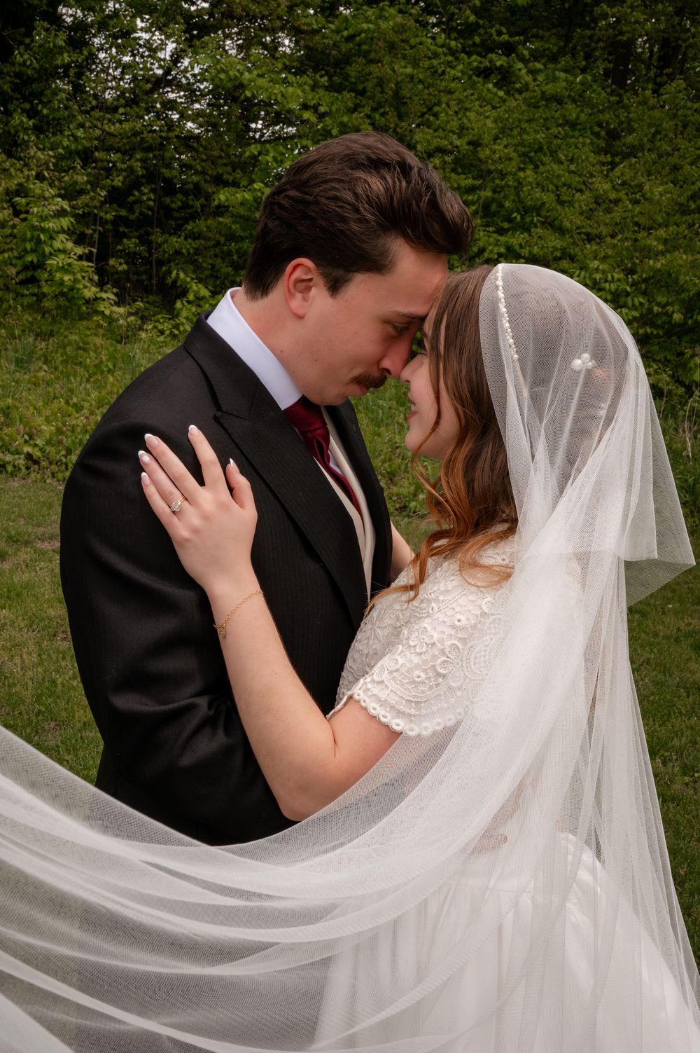 Bride and groom share intimate forehead touch beneath flowing veil in lush green outdoor setting.