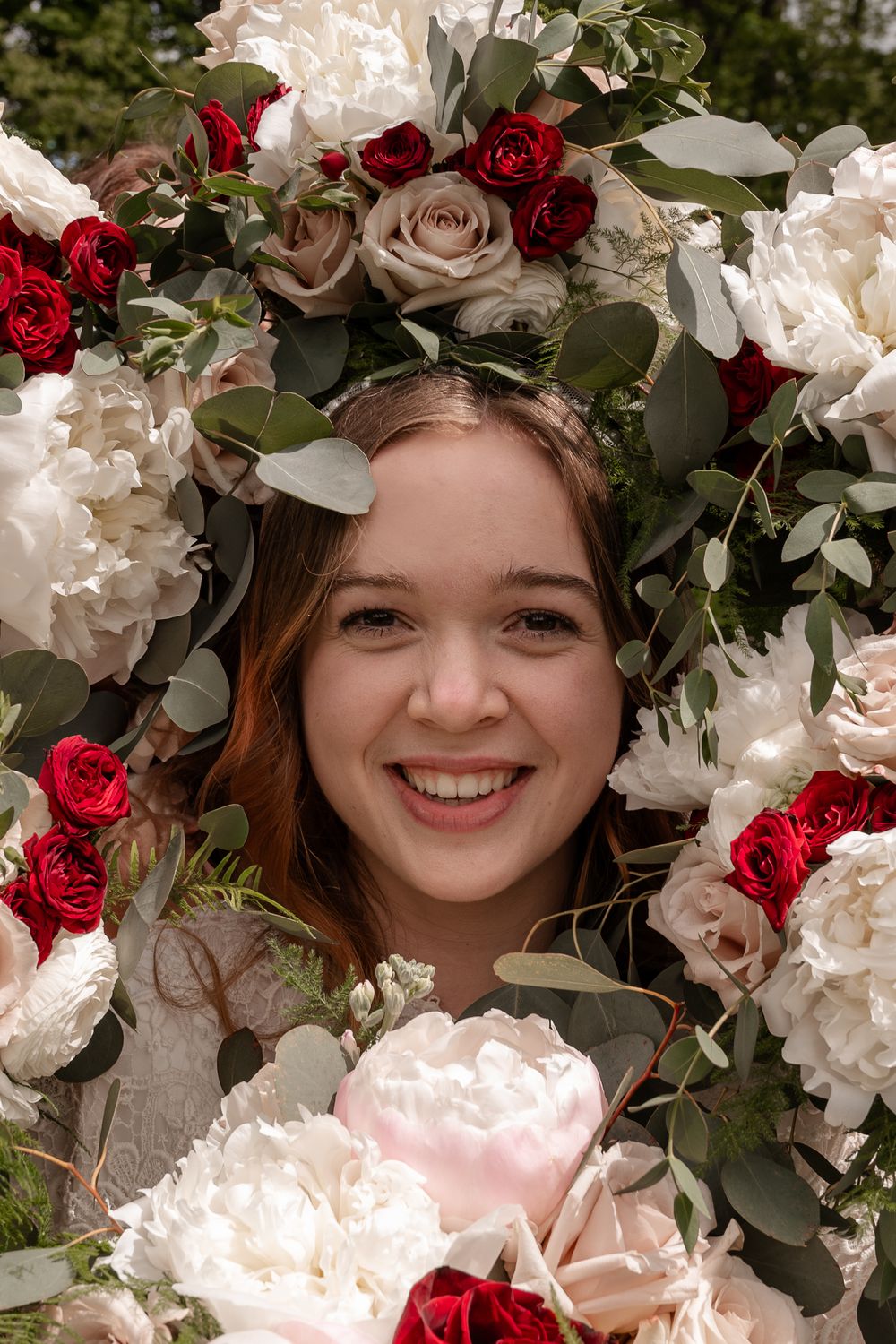 Smiling bride peers through stunning floral arch of white peonies, red roses, and eucalyptus greenery.